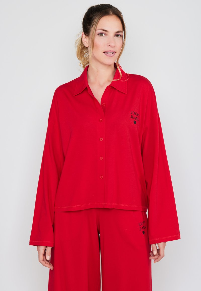 Punainen pitkähihaisen napitettavan paidan, jossa on kaulus ja logo. Yhdistetty tilavien housujen kanssa, jotka ovat molemmat pehmeästä kankaasta ja joissa on näkyvät saumat.