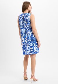 Een vrouw draagt een mouwloze jurk tot de knie met blauw en witte abstracte patronen, gecombineerd met naakte hoge hakken, terwijl ze zijwaarts staat.