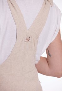 Beige linnen overall met brede bandjes en een V-vormige rug. Klein "cache coeur" label op het bandje, in combinatie met een wit T-shirt eronder.