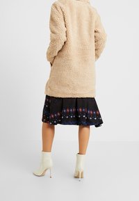 Beige, nadýchané svrchní oblečení s texturovaným povrchem, nošené přes černou sukni s barevným geometrickým vzorem, doplněné krémovými kotníkovými botami.