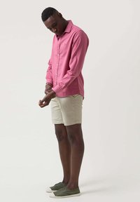 Camisa larga de manga larga en color rosa claro con cierre de botones, pantalones cortos beige y zapatillas de lona verdes. La camisa tiene un cuello y un corte relajado.