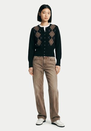 Jeune femme portant un cardigan noir à motif losanges sur une chemise blanche, un jean marron ample et des mocassins blancs, debout devant un fond uni.