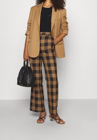 Hellbrauner Blazer über einem schwarzen Oberteil, kombiniert mit karrierten braunen und schwarzen weiten Hosen. Schwarze strukturierte Handtasche und Riemchensandalen.