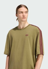 Olivově zelené tričko s krátkými rukávy, s vínovým třípruhovým vzorem na ramenou a jemně vyšitým logem na hrudi. Měkký bavlněný materiál.