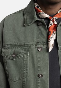 Grøn denimjakke med metalknapper, to brystlommer og spids krave. Et mønstret silkeslips er bundet om halsen.