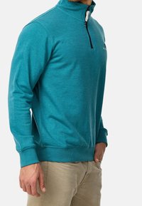 Sudadera de color turquesa con cremallera, cuello alto, puños y bajo acanalados. Fabricada con material suave, presenta un pequeño logo en el pecho.