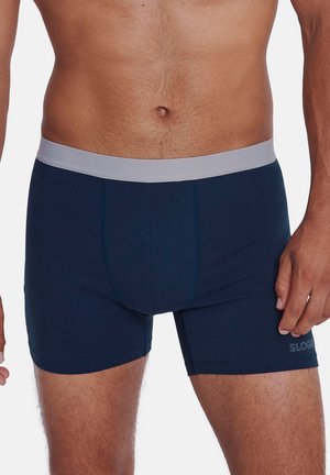 Männlicher Oberkörper, der dunkelblaue Boxershorts mit einem hellgrauen Bund trägt, vor einem schlichten Hintergrund stehend.