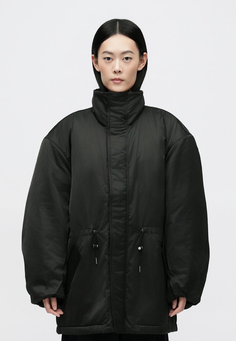 Schwarze gepolsterte Jacke mit hohem Kragen, lockerer Passform und Kordelzug in der Taille. Verfügt über große Taschen und eine glatte Textur. Minimalistisches Design.