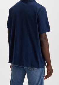 Marinblå kortärmad polo med mjuk textur, avslappnad passform och sidslitsar. Kombinerad med ljusblå denimjeans.