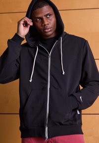 Sudadera negra con capucha, cierre frontal de cremallera, cordones ajustables y bolsillos laterales. Presenta un logo de Nike en la manga.