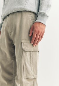Pantalon cargo beige avec poches latérales, en tissu doux. Détails de couture visibles et coupe décontractée. Sweat-shirt gris partiellement visible.