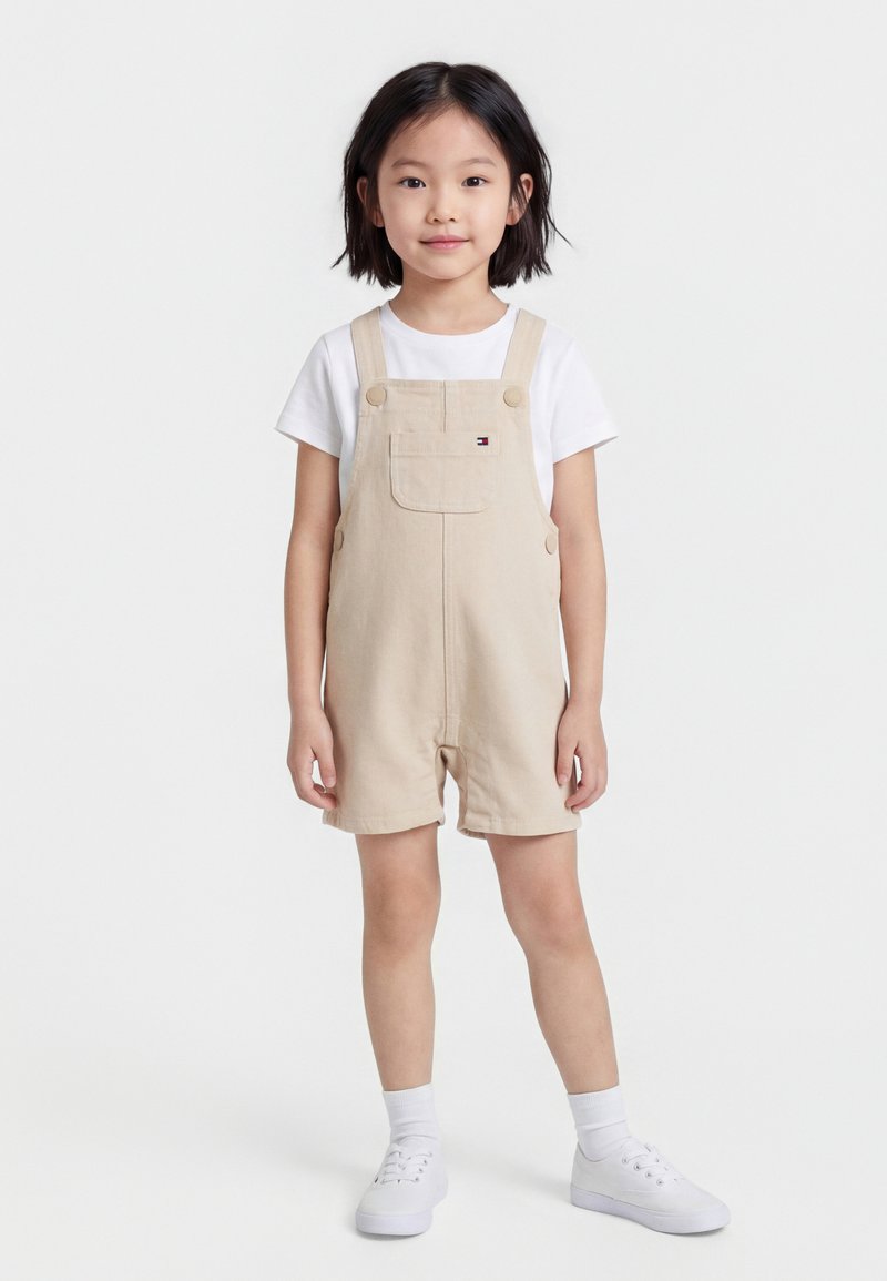 Junges Mädchen steht, trägt beigefarbene kurze Overalls über einem weißen T-Shirt mit weißen Socken und Turnschuhen, vor einem schlichten hellen Hintergrund.