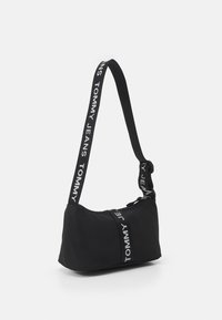 Black shoulder bag with a curved design, zip closure, and adjustable strap featuring white "TOMMY JEANS" text along the length.