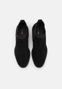 Bottes chelsea noires en suède avec des panneaux latéraux élastiques, surface texturée et détails cousus le long de la semelle. Pas d'accents matériels visibles.