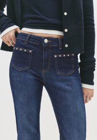 Nő sötétkék farmerben, az elülső zsebein szegecsekkel, sötétkék kardigánban, és gyűrűkkel a bal kezén, a kezei a dereka mellett.