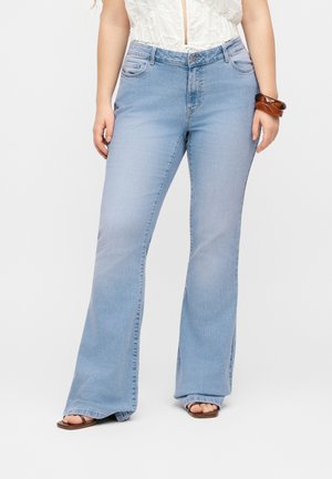 Γυναίκα που φοράει ανοιχτό μπλε καμπάνα τζιν, λευκή μπλούζα με βολάν, καφέ σανδάλια και καφέ βραχιόλια στον δεξί καρπό, στέκεται μπροστά σε λευκό φόντο.