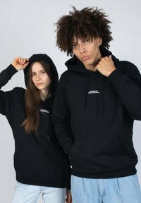 Musta huppari, jossa on etutasku ja rintalogotulostus. Kangas vaikuttaa paksulta ja pehmeältä, ja se on normaalia istuvuutta.