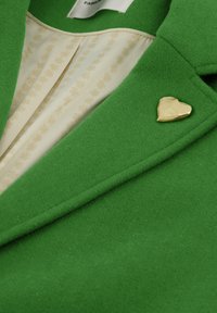 Groene wollen blazer met een hartvormige gouden speld en een crèmekleurige voering met een subtiel bloemenpatroon. Getextureerde stof met gladde accenten.