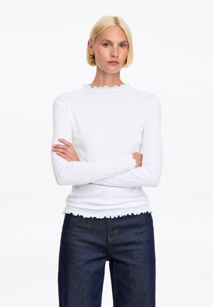 Mujer rubia con el cabello corto, vestida con una camiseta blanca de manga larga y vaqueros azul oscuro, de pie con los brazos cruzados contra un fondo liso.