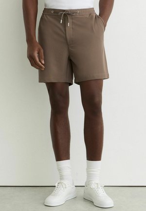 Férfi barna zsinóros rövidnadrágban, fehér bokazokniban, fehér sportcipőben és fehér felsőben, egy egyszínű fal előtt áll.