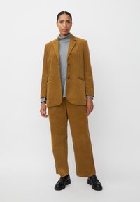 Corduroy blazer en broek set in mosterdgeel, gecombineerd met een grijze coltrui. Heeft twee knopen, zijzakken en een textuurstof.