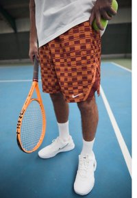 Adulto sosteniendo una raqueta de tenis naranja en una mano y dos pelotas de tenis en la otra, de pie en una pista cubierta de tenis azul.