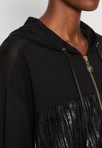Felpa nera con cappuccio e zip, caratterizzata da pieghe di paillettes nere lucide e accenti tattili neri lungo le spalle. Dettagli in hardware dorato.