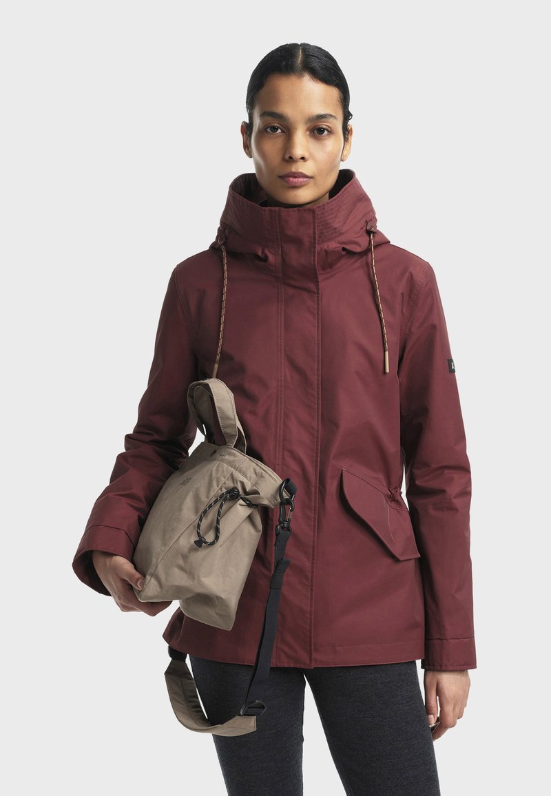 Rode waterdichte jas met hoge kraag, verstelbare koordjes en grote zijzakken. Beige trekkoordtas met zwarte strap in de hand gehouden.