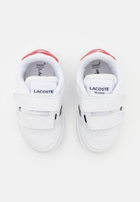 Pár bielych detských tenisiek Lacoste s suchými zipsami a červenými akcentmi, pohľad zhora na bielom pozadí.