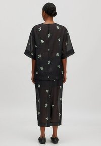 Schwarzes durchscheinendes Outfit aus Stoff, bestehend aus einem lockeren Oberteil und einem passenden Rock mit floralen Stickereien in Weiß und Grün. Kurze Ärmel und knielange Länge.