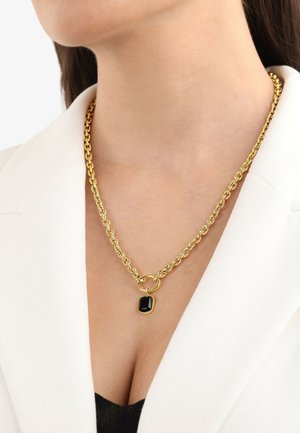 Collana in oro con maglie circolari intrecciate e un pendente rettangolare nero, posizionata su una blusa chiara.