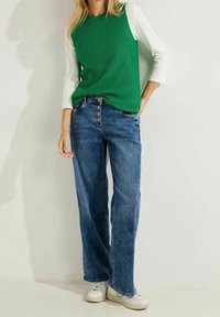 Pull en tricot côtelé vert avec manches blanches, associé à un jean large bleu clair avec braguette boutonnée et des baskets blanches.