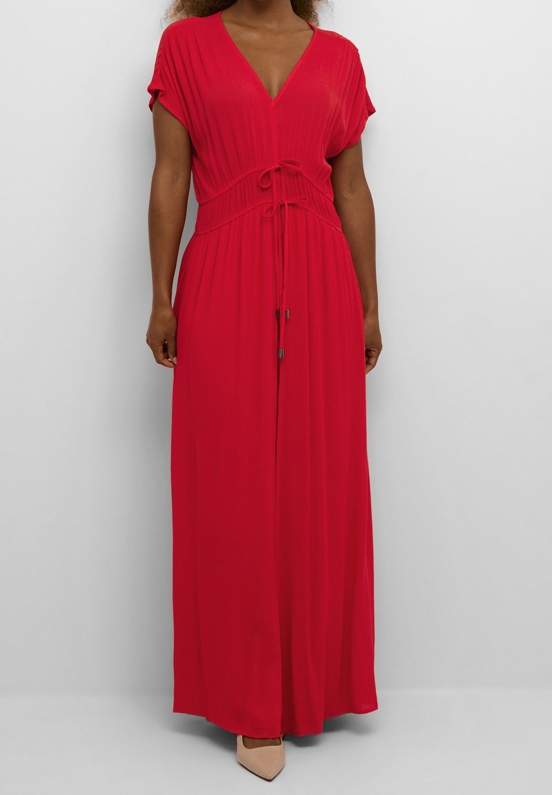 Femme portant une robe rouge longue jusqu'au sol avec un col en V, des manches courtes et deux liens à la taille, associée à des talons beiges pointus.