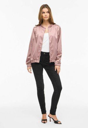 Kvinde iført en lyserød satin bomberjakke, hvid top, sorte skinny jeans og sorte åben-tå hælede sandaler, stående mod hvid baggrund.