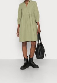 Robe vert clair, longueur genou, avec un col, des manches 3/4 et une taille froncée. Portée avec des bottes noires à la cheville et un sac à main noir.