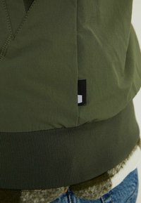 Gros plan d'une veste vert olive avec ourlet côtelé, montrant une petite étiquette rectangulaire noire et blanche sur le côté au-dessus d’un jean en denim bleu.