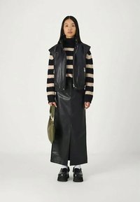 Γυναίκα φορώντας μια μαύρη δερμάτινη γιλέκα και φούστα, ρίγες μαύρου και μπεζ πουλόβερ, κρατώντας μια ελαιόχρωμη τσάντα, standing σε λευκό φόντο.