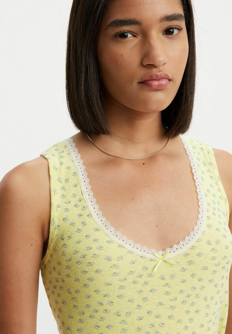 Top sin mangas amarillo con patrón floral, cuello con puntilla y una cinta decorativa en el centro. Hecho de una tela suave y texturizada.