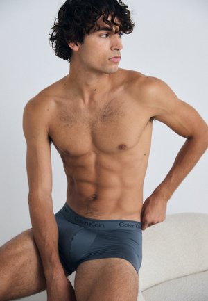 Grijze katoenen slips met een laag uitgesneden ontwerp, voorzien van een zachte tailleband met het logo "Calvin Klein". Het model heeft een getraind figuur en krullend haar.