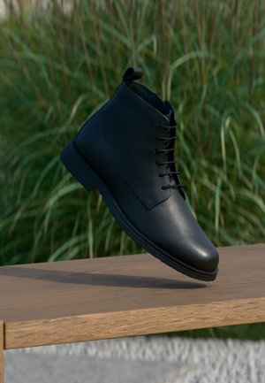Schwarzer Leder-Stiefelette mit Schnürsenkeln, schwebend über einer Holzbank mit grünen Pflanzen im Hintergrund.