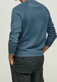 Muž v modrém pleteném svetr s dlouhým rukávem a černých kalhotách, se založenýma rukama v kapsách, zobrazený ze zadu na jednoduším pozadí.