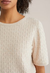 Béžový pletený sveter s geometrickým vzorom, krátkymi rukávmi a rebrovaným výstrihom. Mäkká tkanina s jemným leskom.