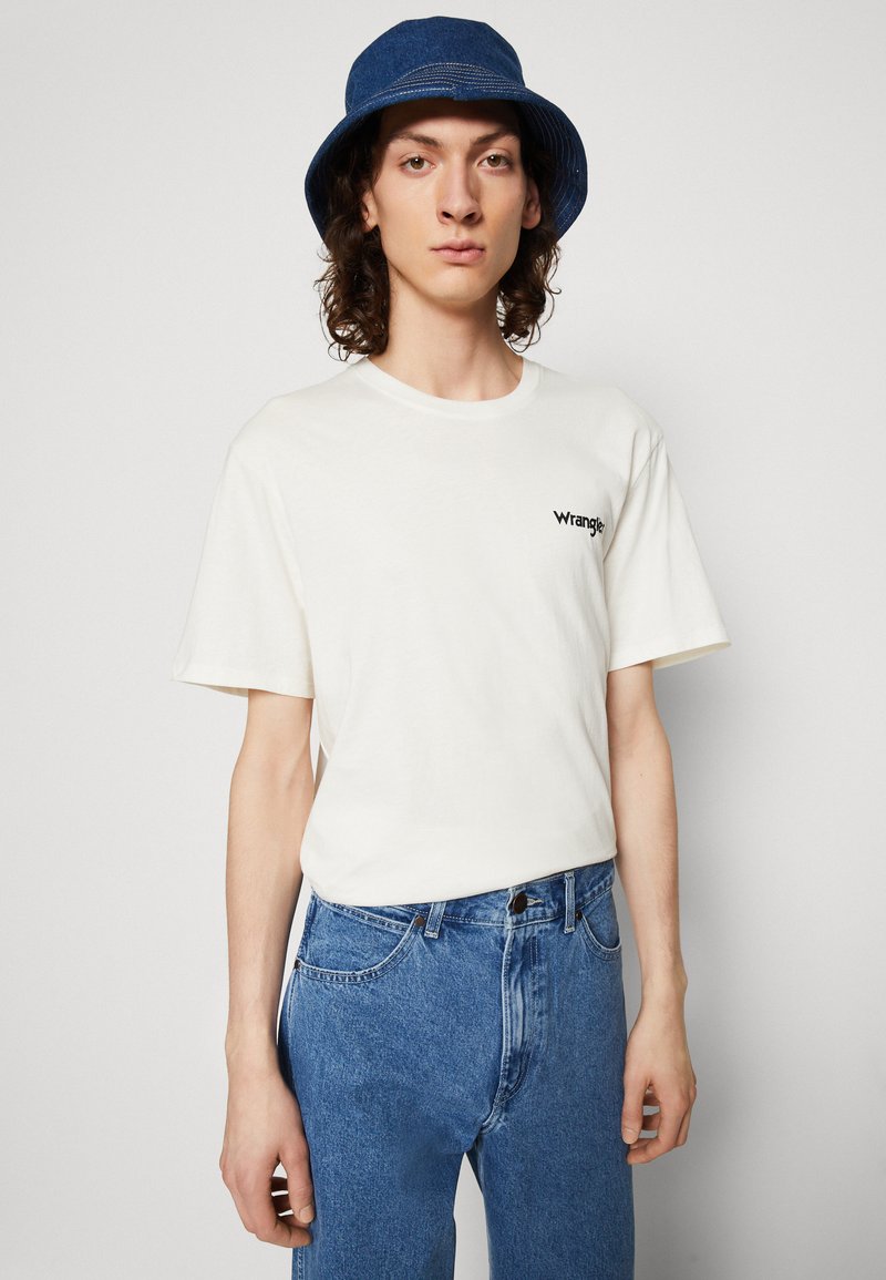 T-shirt bianco in cotone con maniche corte e logo "Wrangler" nero. Abbinato a jeans in denim blu e un cappello a secchiello blu. Vestibilità casual.