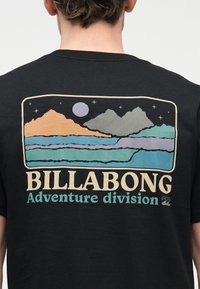 Πίσω μέρος μαύρου πουκαμίσου με πολύχρωμο γραφικό βουνού και λίμνης κάτω από νυχτερινό ουρανό και κείμενο "BILLABONG Adventure division."