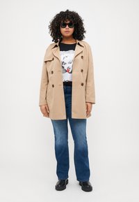 Beige trenchcoat med dobbelspent design, store knapper og krage. Kledd med en svart grafisk T-skjorte, blå jeans og svarte støvler.