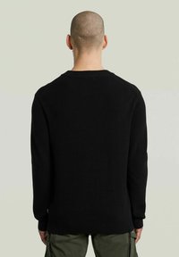 Maglione nero a maglia con texture a coste, scollo rotondo e maniche lunghe. Presenta una vestibilità comoda ed è abbinato a pantaloni verde oliva.