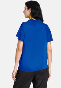 Kungsblå kortärmad blus med en slät struktur och krage, i kombination med svarta byxor. Har en avslappnad passform och subtila draperingar.