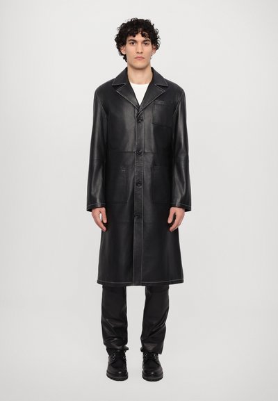 Sort læderfrakke med krave, frontknapper, sidelommer og synlige syninger. Slim fit, båret med matchende sorte bukser.