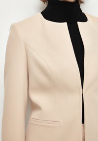 Beige texturerad blazer med en minimalistisk, kraglös design och en sidoficka, lager på en svart rullekragströja.