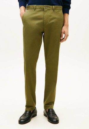 Mężczyzna w oliwkowozielonych, dopasowanych spodniach, z rękami w kieszeniach, w czarnych skórzanych mokasynach, stojący na tle jednolitego jasnego tła.